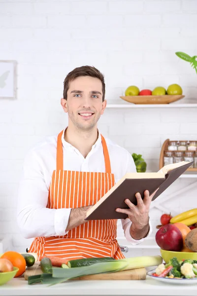 Man with recipe book in hands at table with different products and utensil in kitchen on white wall background