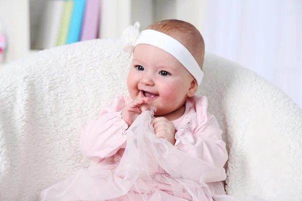 Cute baby girl in pink dress sitting in arm-chair, on home interior background