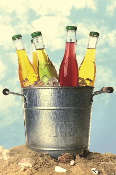 Bottles of tasty drink in metal bucket with ice on sand isolated on white