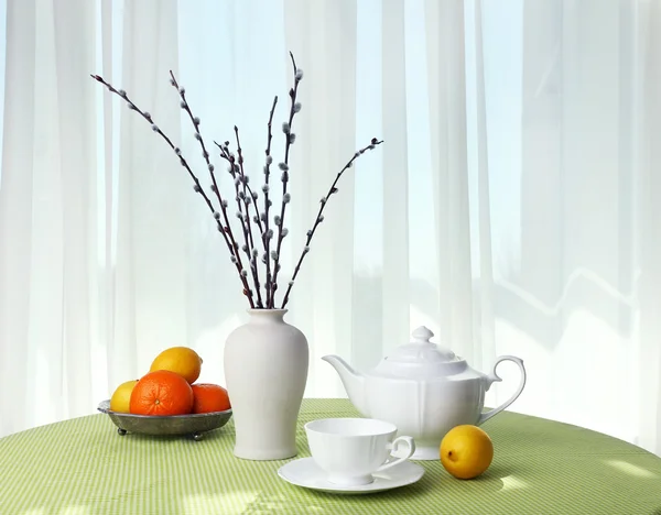 Willow twigs, teapot, cups and citrus on table on curtains background