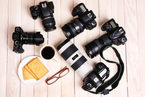 Modern cameras with glasses, cup of coffee and cookies on wooden table, top view