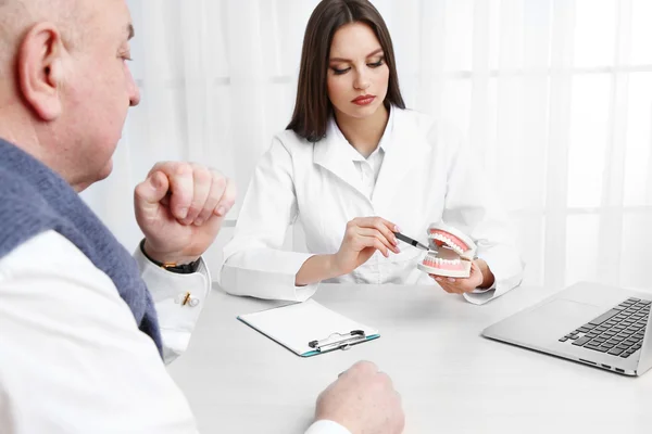 Young female dentist showing model of human teeth in her office at wooden table on white curtain background