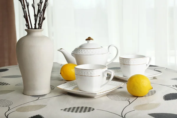 Willow twigs in vase with teapot, cups and lemon on table on curtains background