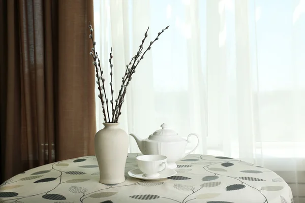 Willow twigs in vase with teapot and cup on table on curtains background