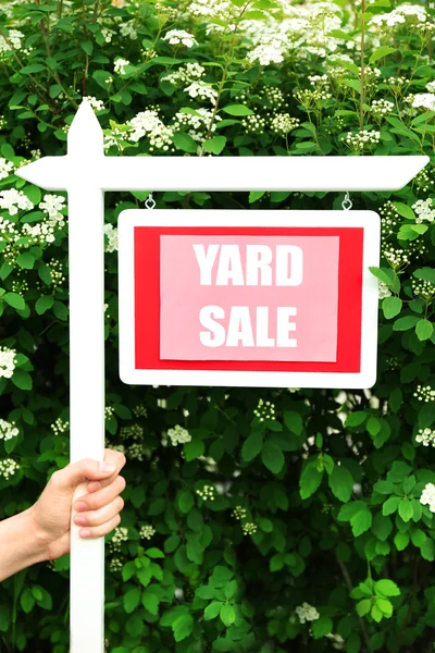 Wooden Yard Sale sign in female hand over green bush and flowers background