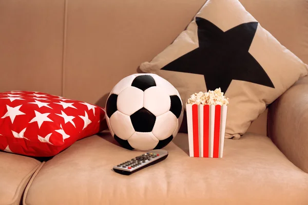 Soccer ball, remote control and box of popcorn on comfortable sofa, indoors