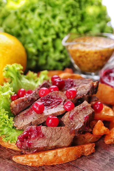 Beef with cranberry sauce, roasted potato slices and bun on wooden cutting board, close-up