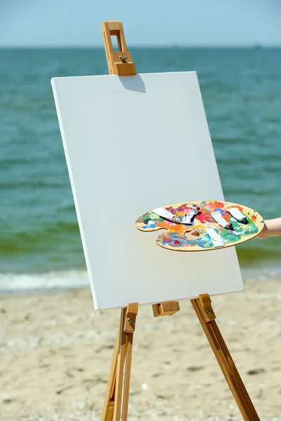 Palette with paints and easel with canvas on beach