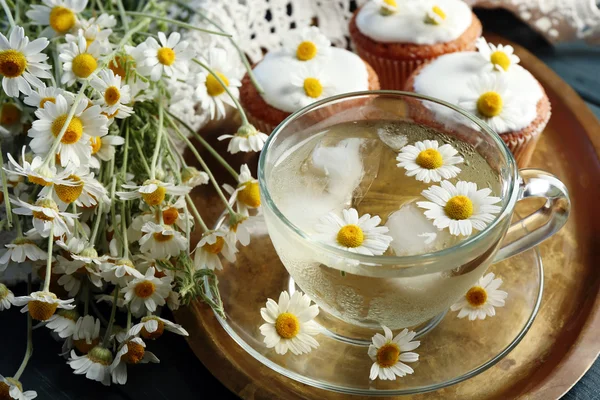 Cup of chamomile tea with chamomile flowers and tasty muffins on tray, on color wooden background