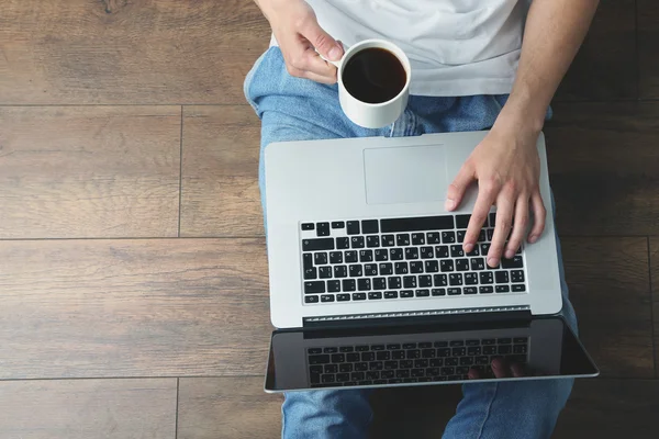 Young man sitting on floor with laptop and cup of coffee in room