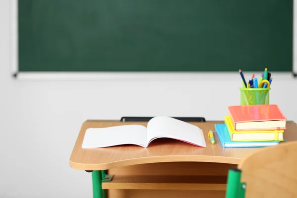 Wooden desk with stationery and chair in class on blackboard background