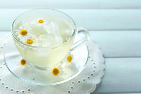 Glass of cold chamomile tea with ice cubes and chamomile flowers on color wooden background