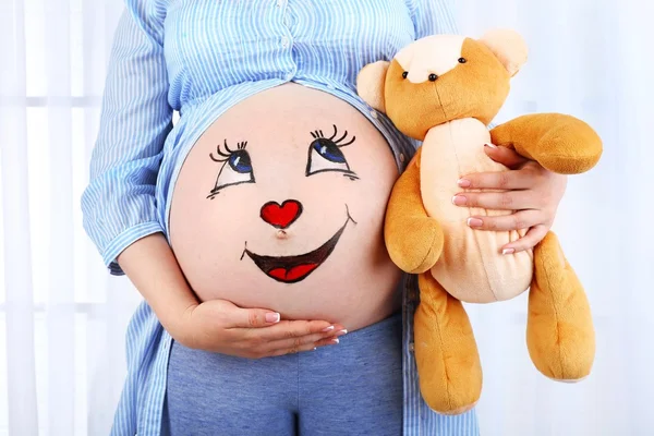 Beautiful young pregnant with baby toy and picture on her belly, on light background