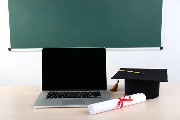 Notebook, diploma and master hat on desk in class on blackboard background