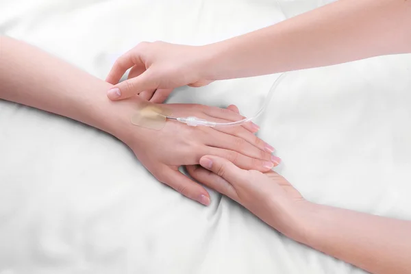Female hands holding patient hand with dropper needle on bed close-up