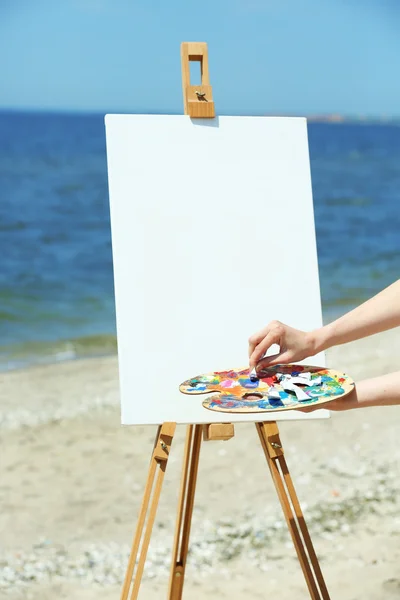 Female hand holding palette with paints and easel with canvas on beach