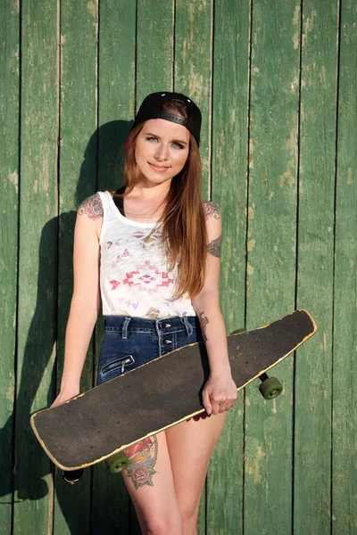 Beautiful girl with tattooed body, holding skateboard on wooden wall background