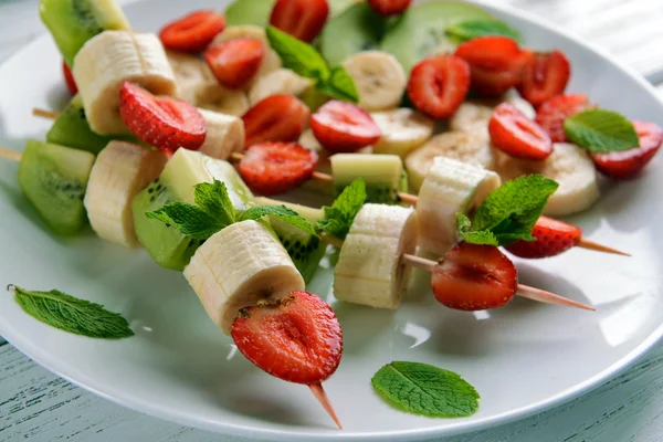 Fresh fruits on skewers in plate on wooden table, closeup