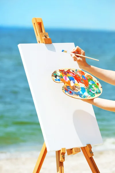 Female hand holding palette with paints and easel with canvas on beach