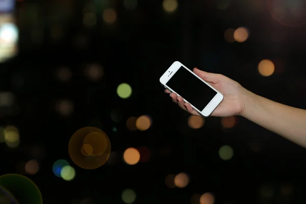 Female hand holding touch screen mobile phone on blurred night lights background