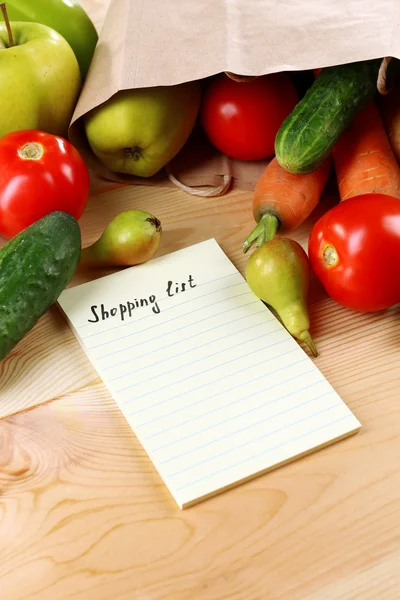 Heap of fruits and vegetables with shopping list on table close up