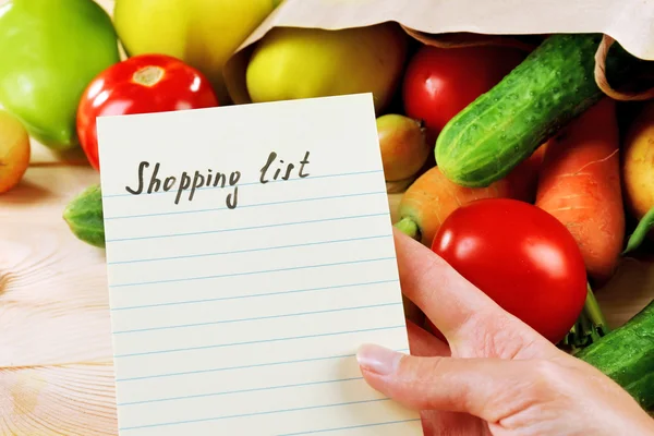Heap of fruits and vegetables with shopping list on table close up