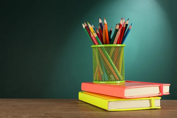 Books and metal holder of crayons on desk on green chalkboard background
