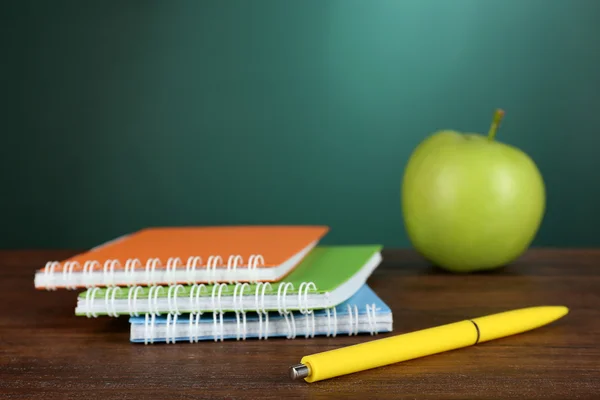 Colorful notebooks with pen and green apple on desk on green chalkboard background