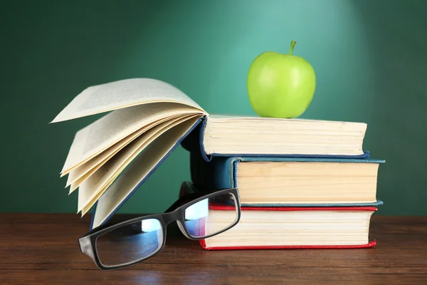 Stack of books, glasses and green apple on desk on green chalkboard background