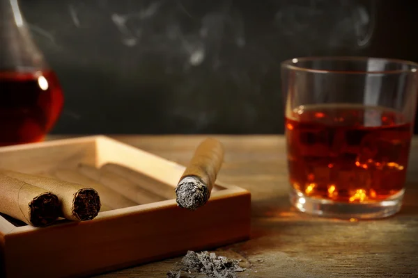 Cigars and burning one with cognac on wooden table on black background
