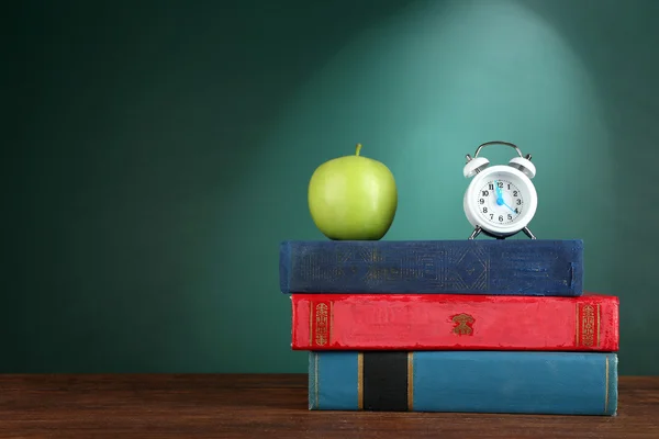 Stack of books with alarm clock and green apple on green chalkboard background