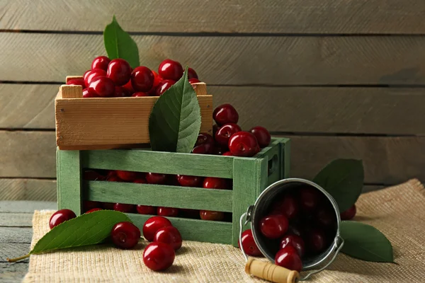 Sweet cherries with green leaves in bucket and wooden boxes, on wooden background
