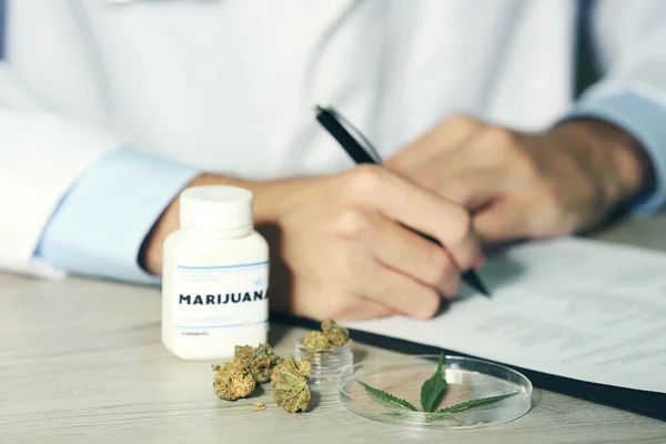 Doctor writing on prescription blank and bottle with medical cannabis on table close up