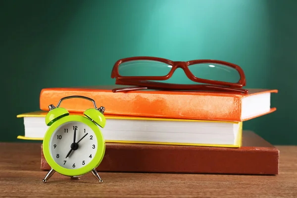 Stack of books with glasses and alarm clock on desk on green chalkboard background