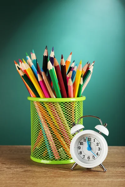 Metal holder with crayons near alarm clock on desk on green chalkboard background