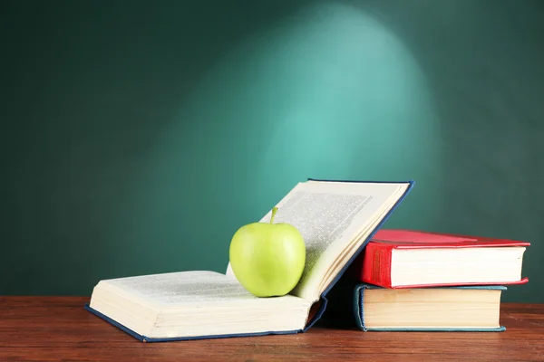 Open books and green apple on desk on green chalkboard background