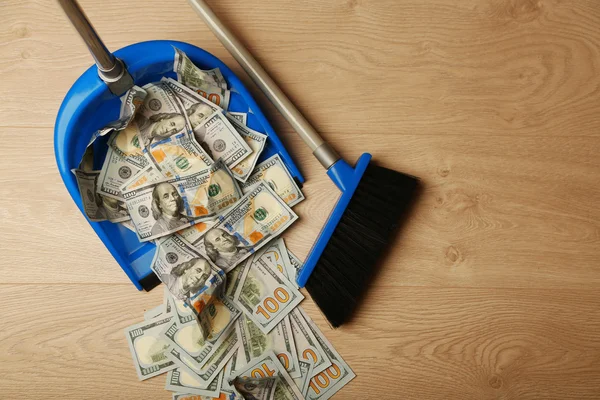 Broom sweeps dollars in garbage scoop on wooden floor background