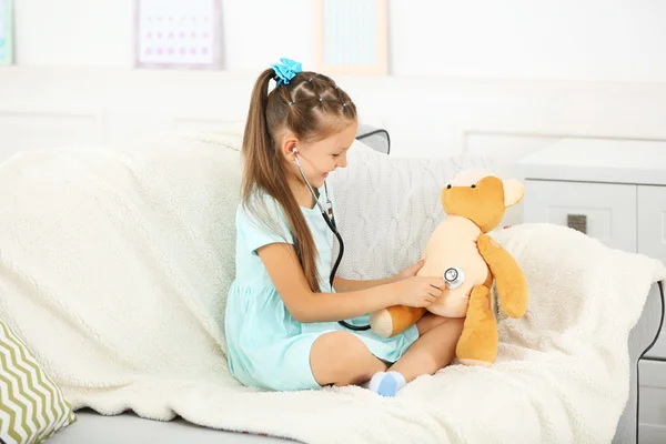 Beautiful little girl with teddy bear sitting on sofa, on home interior background