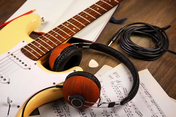 Electric guitar with headphones, musical notes and note book on wooden background