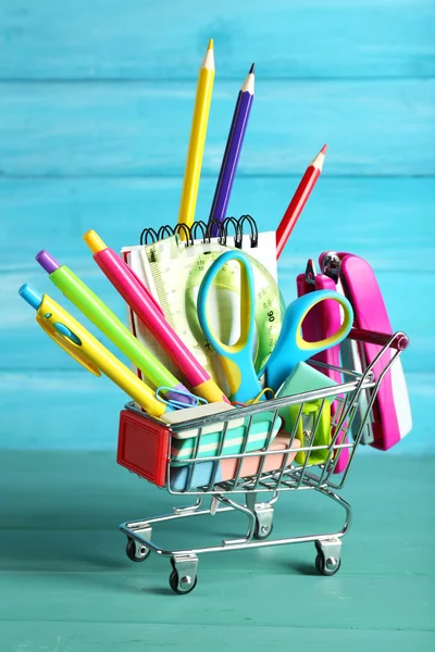 Bright stationery objects in mini supermarket cart on blue wooden background