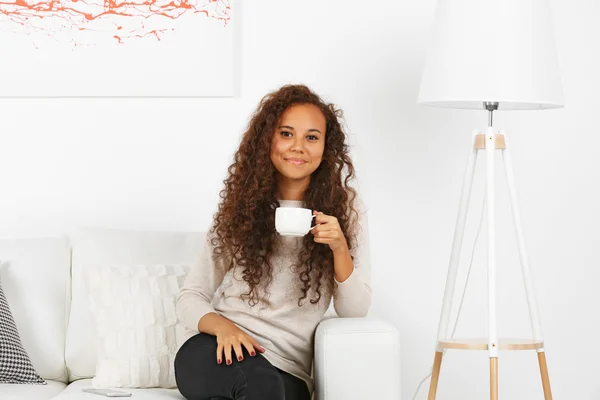 Young woman sitting on comfortable sofa with cup on coffee in hands in white room