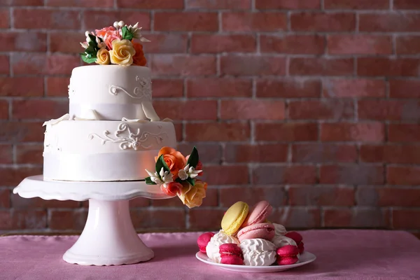 Beautiful wedding cake decorated with flowers and plate with cakes on pink table against brick wall background