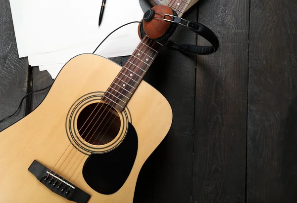 Acoustic guitar, headphones, musical notes and white papers on wooden background, close up