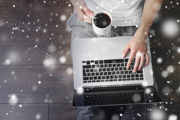 Young man sitting on floor with laptop and cup of coffee over snow effect