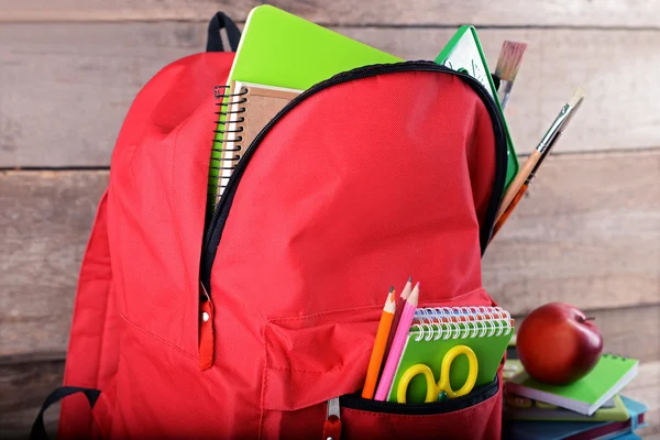 Full of stationary red backpack and pile of books with apple on top on wooden background
