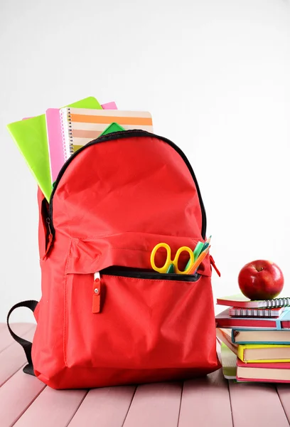 Full of stationary red backpack and pile of books with apple on top on wooden table against white background