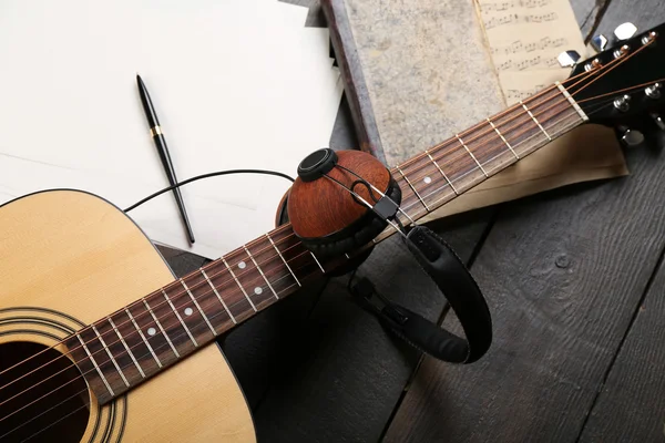 Acoustic guitar, headphones, musical notes and white papers on wooden background, close up