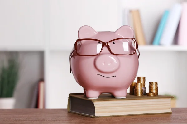 Piggy bank in glasses with book and coins on home or office background