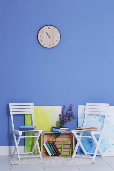 Room design with white chairs, bookcase, pictures, flowers and clock over blue wall