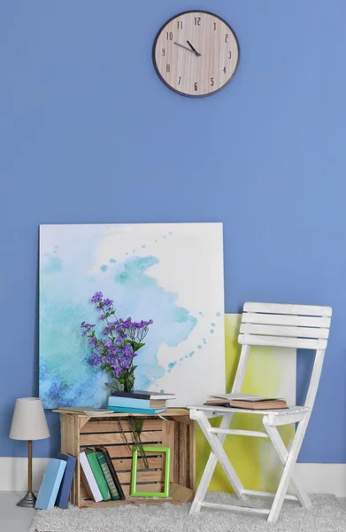 Room design with white chair, bookcase, pictures, lamp, flowers and clock over blue wall